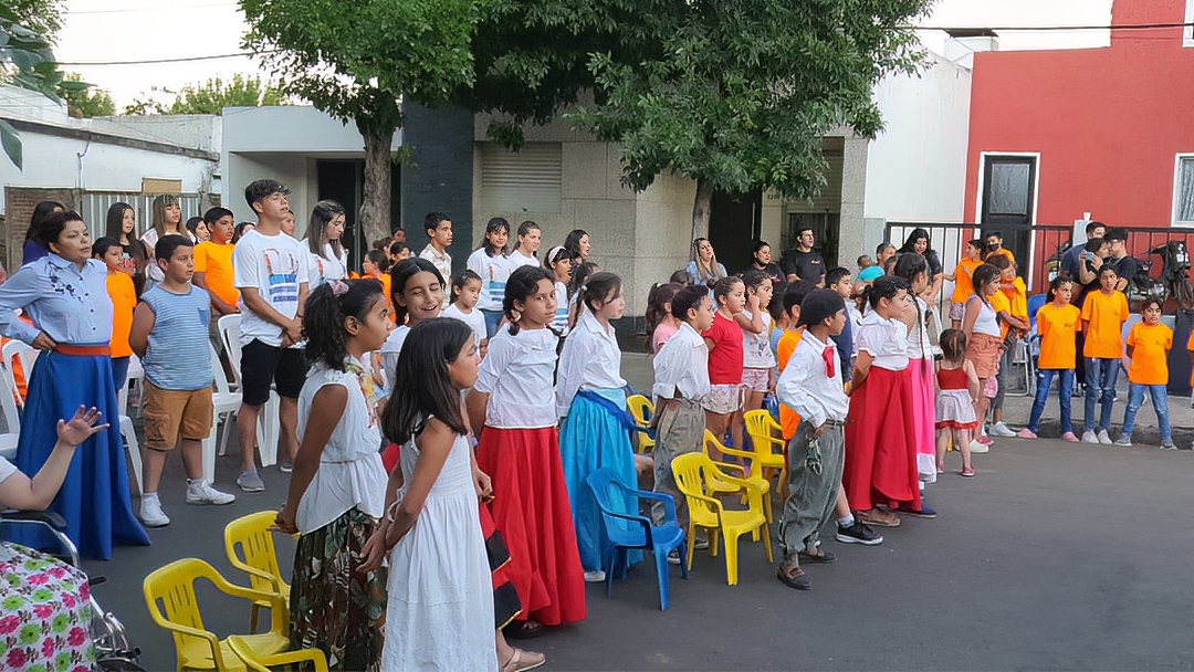 Children in colorful skirts and white tops and orange t-shirts standing in an outdoor courtyard in a semicircle. They each have a chair behind them and a row of adults is behind them also standing.