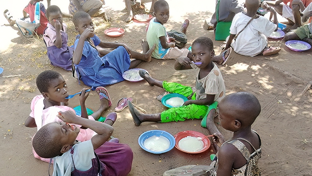 Kids sitting on ground eating food from bowls with spoons